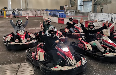 A group of five people in helmets and masks sit in red and white go-karts indoors, raising their hands and posing before a race.