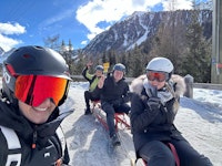 Four people in winter gear and helmets sit on sleds in the snow, smiling and posing for the camera with snowy mountains and trees in the background.