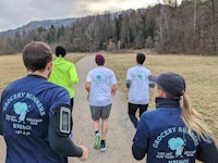 Four people jog on a gravel path in a park, wearing "Grocery Runners" shirts; trees and hills are visible in the background.