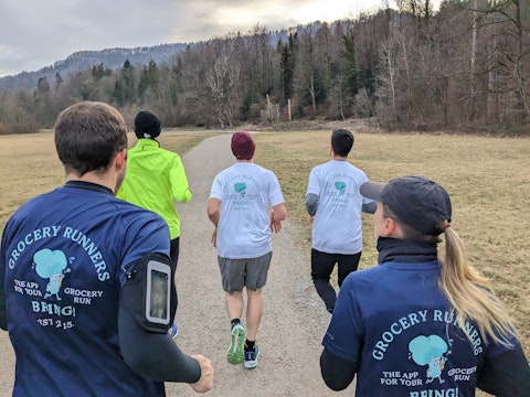 Four people jog on a gravel path in a park, wearing "Grocery Runners" shirts; trees and hills are visible in the background.