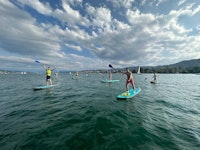 A group of people paddleboarding on a lake under a partly cloudy sky, with distant hills and sailboats visible in the background.