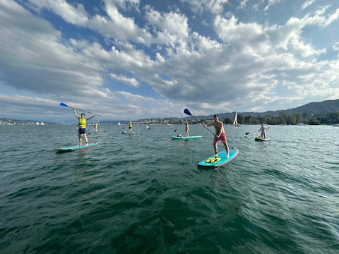 A group of people paddleboarding on a lake under a partly cloudy sky, with distant hills and sailboats visible in the background.