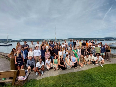 A large group of people pose for a photo outdoors by a lake with sailboats and hills in the background on a partly cloudy day.