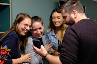 Four people stand close together, smiling and looking at a smartphone, in a casual indoor setting.