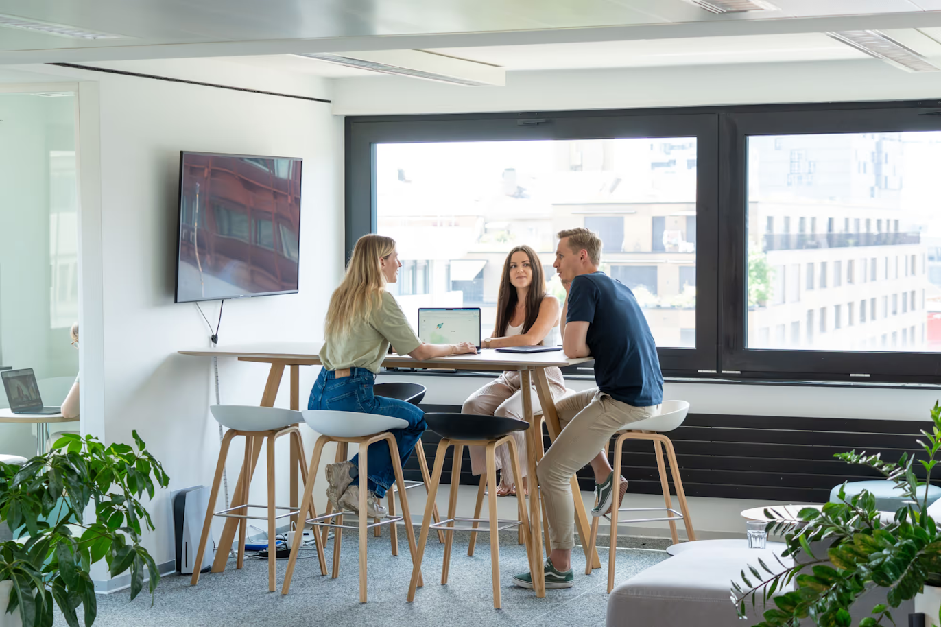 Drei Personen sitzen an einem hohen Tisch in einem modernen Büro und führen eine Diskussion, während vor ihnen ein Laptop geöffnet ist.