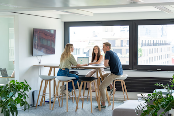Drei Personen sitzen an einem hohen Tisch in einem modernen Büro und führen eine Diskussion, während vor ihnen ein Laptop geöffnet ist.