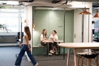 Two women sit at a small table with a laptop in a glass meeting room, while another woman walks by in an open office space.