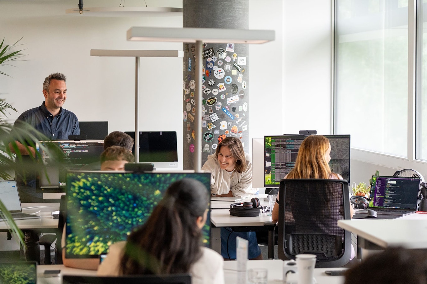 Open-plan office with several people working at desks with computers, one person standing and others sitting, engaged in conversation. Bright space with large windows and decorated pillar.