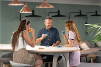 Three people sit around a table in a modern, well-lit room, smiling and drinking from coffee cups.