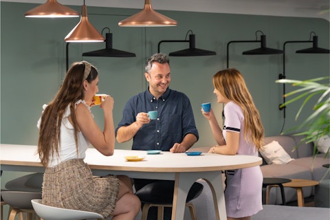Three people sit around a table in a modern, well-lit room, smiling and drinking from coffee cups.