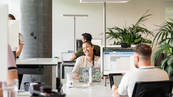 Three people work at desks with computers in a modern, open-plan office space. One person in the center smiles at the camera. Laptops and office plants are visible.