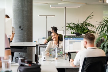 Drei Personen arbeiten an Schreibtischen mit Computern in einem modernen, offenen Büro. Eine Person in der Mitte lächelt in die Kamera. Laptops und Büropflanzen sind zu sehen.