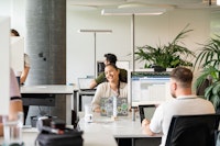 Three people work at desks with computers in a modern, open-plan office space. One person in the center smiles at the camera. Laptops and office plants are visible.