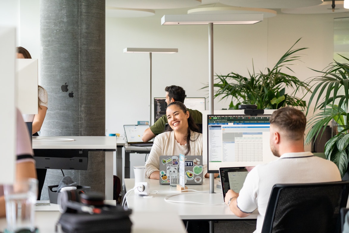 Drei Personen arbeiten an Schreibtischen mit Computern in einem modernen, offenen Büro. Eine Person in der Mitte lächelt in die Kamera. Laptops und Büropflanzen sind zu sehen.
