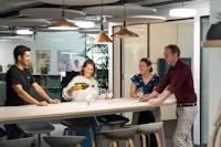 Four people stand around a high table in an office while one person pours wine into glasses; they appear to be enjoying a casual gathering.