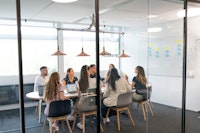 Eight people sit around a table in a glass-walled office meeting room, engaged in discussion, with laptops and notes in front of them.