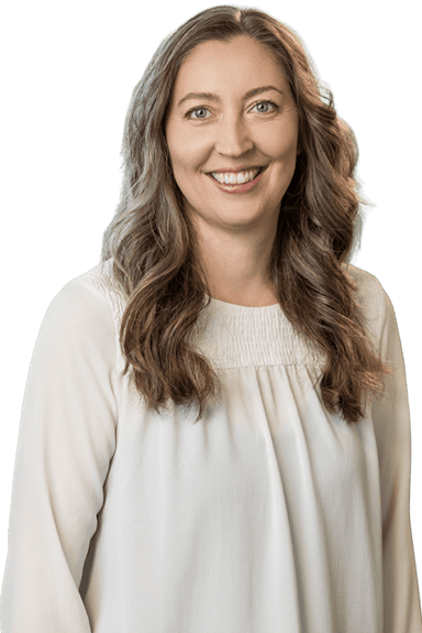 A woman with long wavy brown hair, wearing a white blouse, smiles at the camera against a plain background.