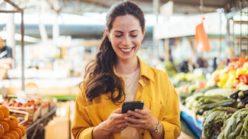 Eine Frau in einer gelben Jacke lächelt, während sie ihr Smartphone in einem Markt benutzt, in dem um sie herum frisches Obst und Gemüse ausgestellt ist.