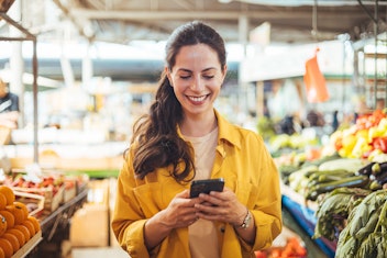 Eine Frau in einer gelben Jacke lächelt, während sie ihr Smartphone in einem Markt benutzt, in dem um sie herum frisches Obst und Gemüse ausgestellt ist.