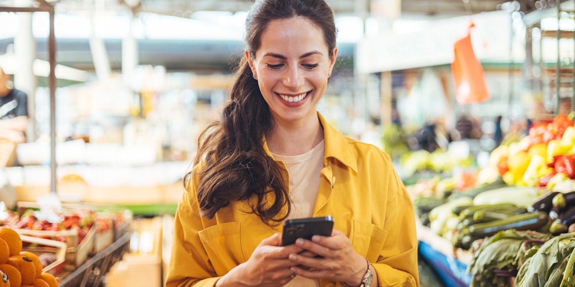 Eine Frau in einer gelben Jacke lächelt, während sie ihr Smartphone in einem Markt benutzt, in dem um sie herum frisches Obst und Gemüse ausgestellt ist.