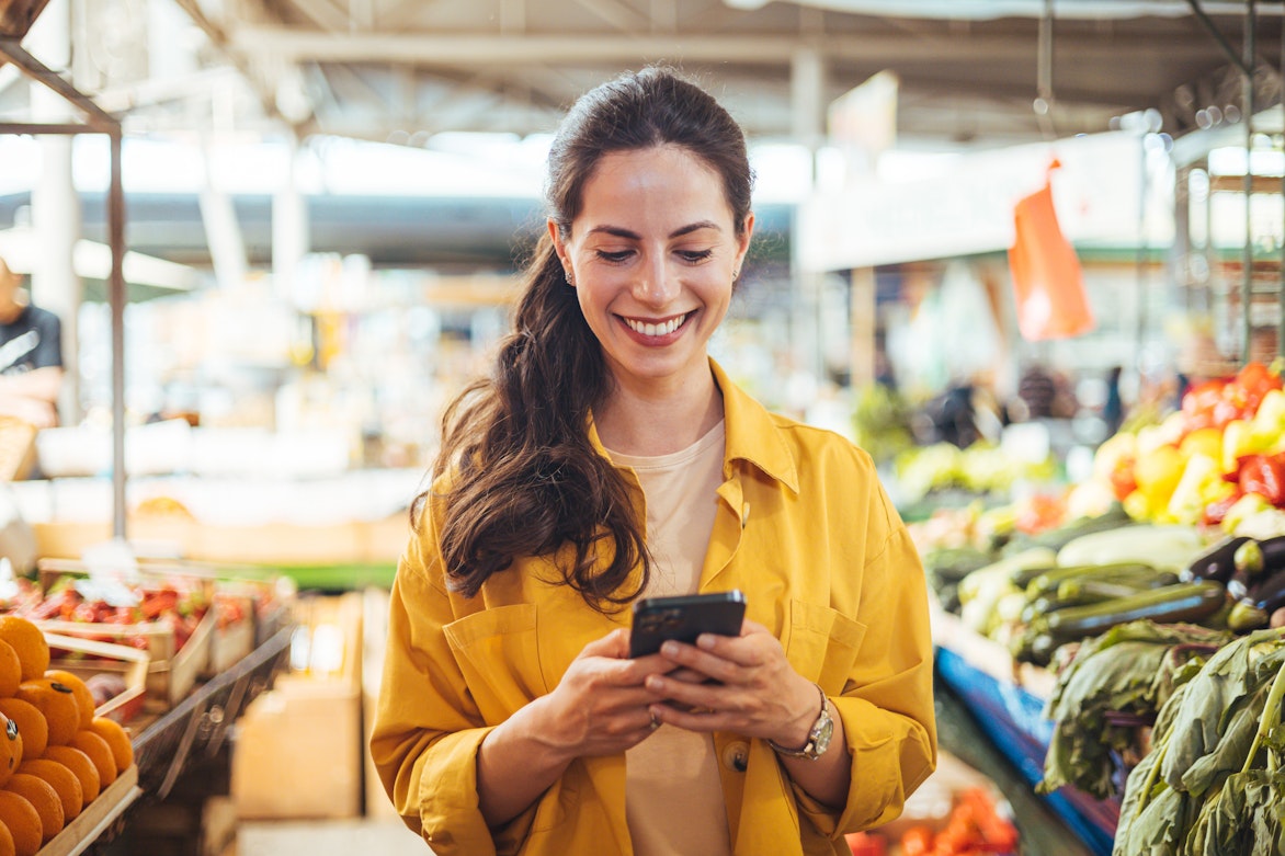 Eine Frau in einer gelben Jacke lächelt, während sie ihr Smartphone in einem Markt benutzt, in dem um sie herum frisches Obst und Gemüse ausgestellt ist.