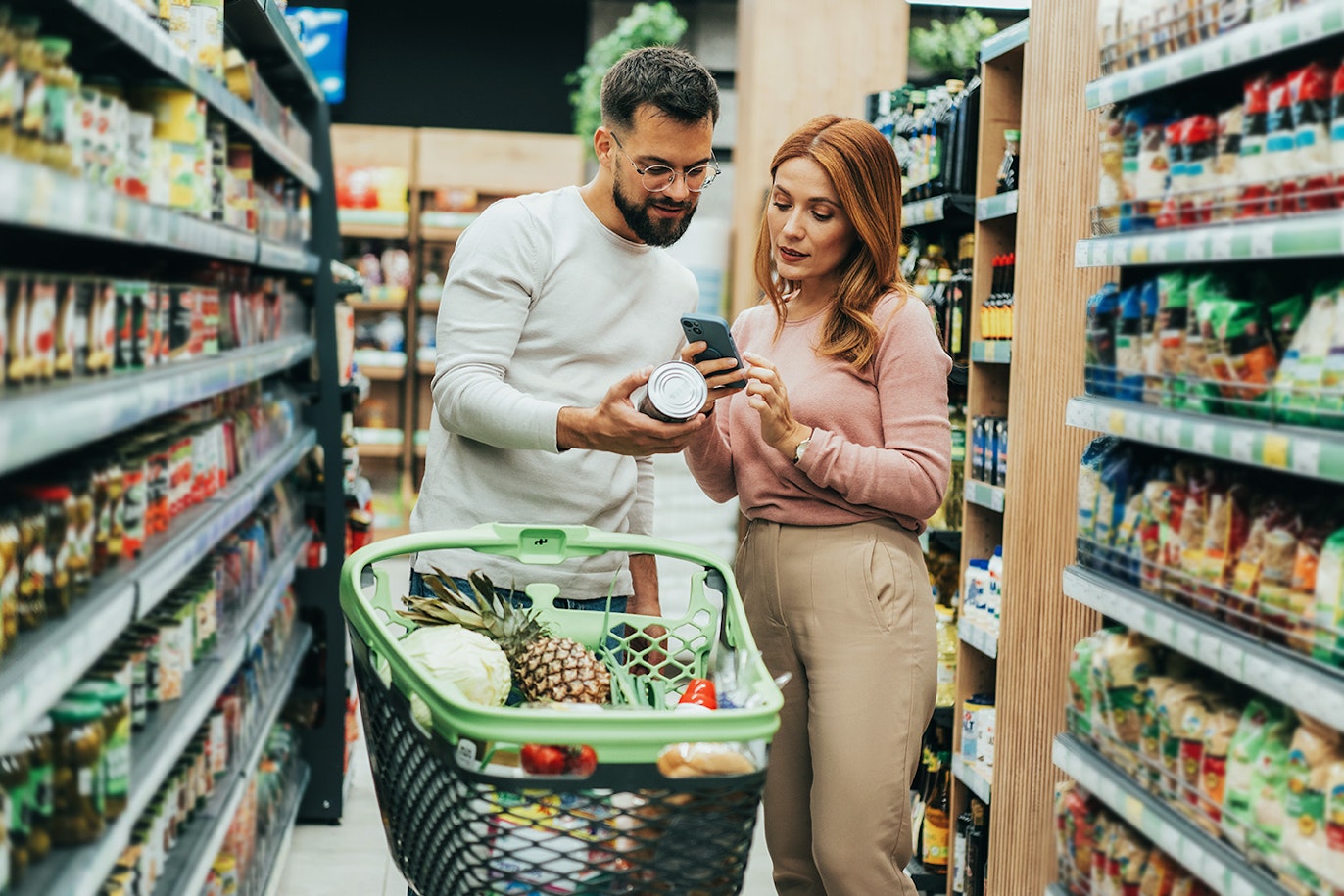 Ein Mann und eine Frau stehen in einem Supermarktgang und betrachten eine Dose und ein Telefon, vor ihnen steht ein Einkaufswagen voller Lebensmittel.