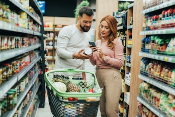 Ein Mann und eine Frau stehen in einem Supermarktgang und betrachten eine Dose und ein Telefon, vor ihnen steht ein Einkaufswagen voller Lebensmittel.