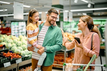 Ein Mann mit einem kleinen Mädchen steht neben Obst und Gemüse in einem Lebensmittelgeschäft, während eine Frau mit einem Smartphone in der Nähe eines Einkaufswagens mit ihnen spricht.