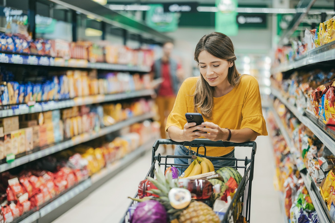 Eine Frau in einem gelben Shirt schiebt einen mit Lebensmitteln gefüllten Einkaufswagen durch einen Supermarktgang und schaut dabei auf ihr Handy.