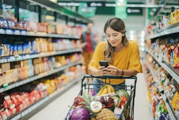 Eine Frau in einem gelben Shirt schiebt einen mit Lebensmitteln gefüllten Einkaufswagen durch einen Supermarktgang und schaut dabei auf ihr Handy.