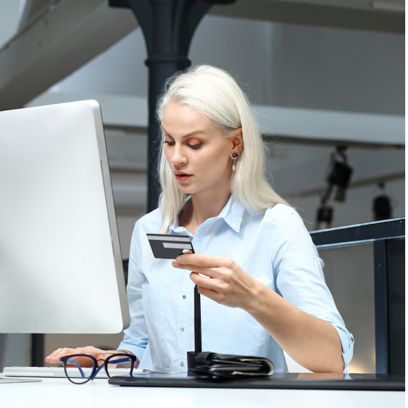 Kobieta w koszuli siedząca przy komputerze i trzymająca kartę płatniczą i pisząca coś na klawiaturze.