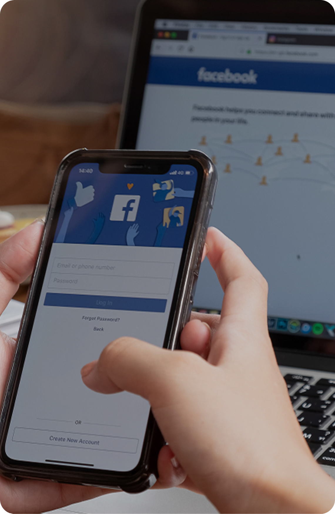 Smartfon z ekranem logowania Facebooka trzymany w dłoni na tle laptopa – symbol prowadzenia kampanii reklamowych Meta Ads i marketingu w social media.