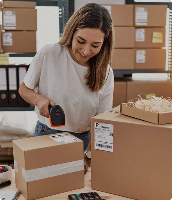 Kobieta przygotowuje paczki do wysyłki i skanuje etykietę na kartonowym pudełku. Na stole stoją zapakowane przesyłki, a w tle widać regały z kolejnymi kartonami i materiałami do obsługi zamówień.