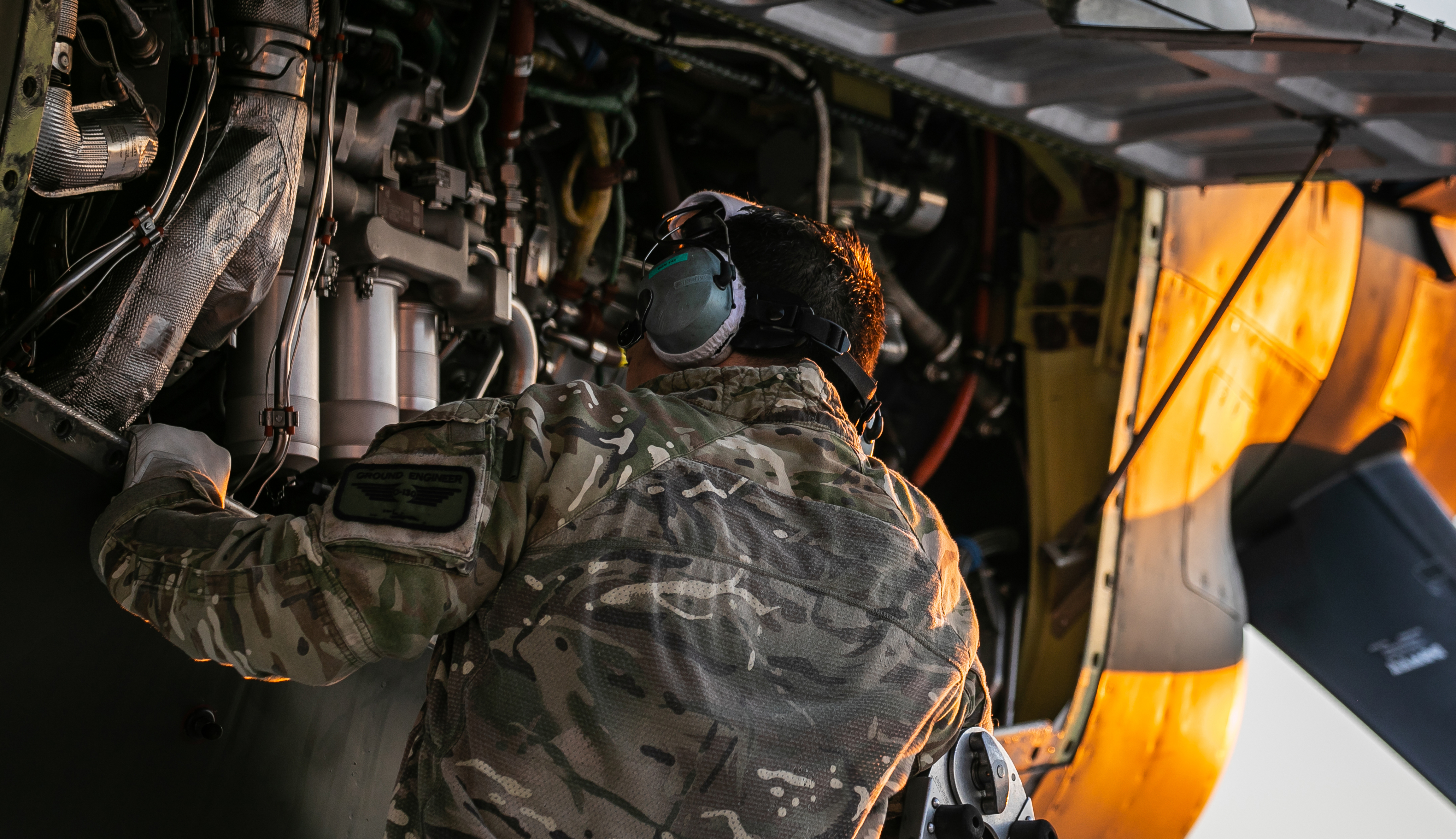 A person in military camouflage uniform inspects or repairs the engine area of an aircraft, with mechanical components visible.