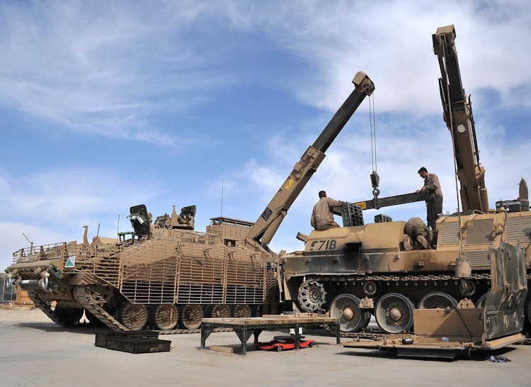 Soldiers conducting maintenance on an armoured vehicle using cranes in a military repair area.