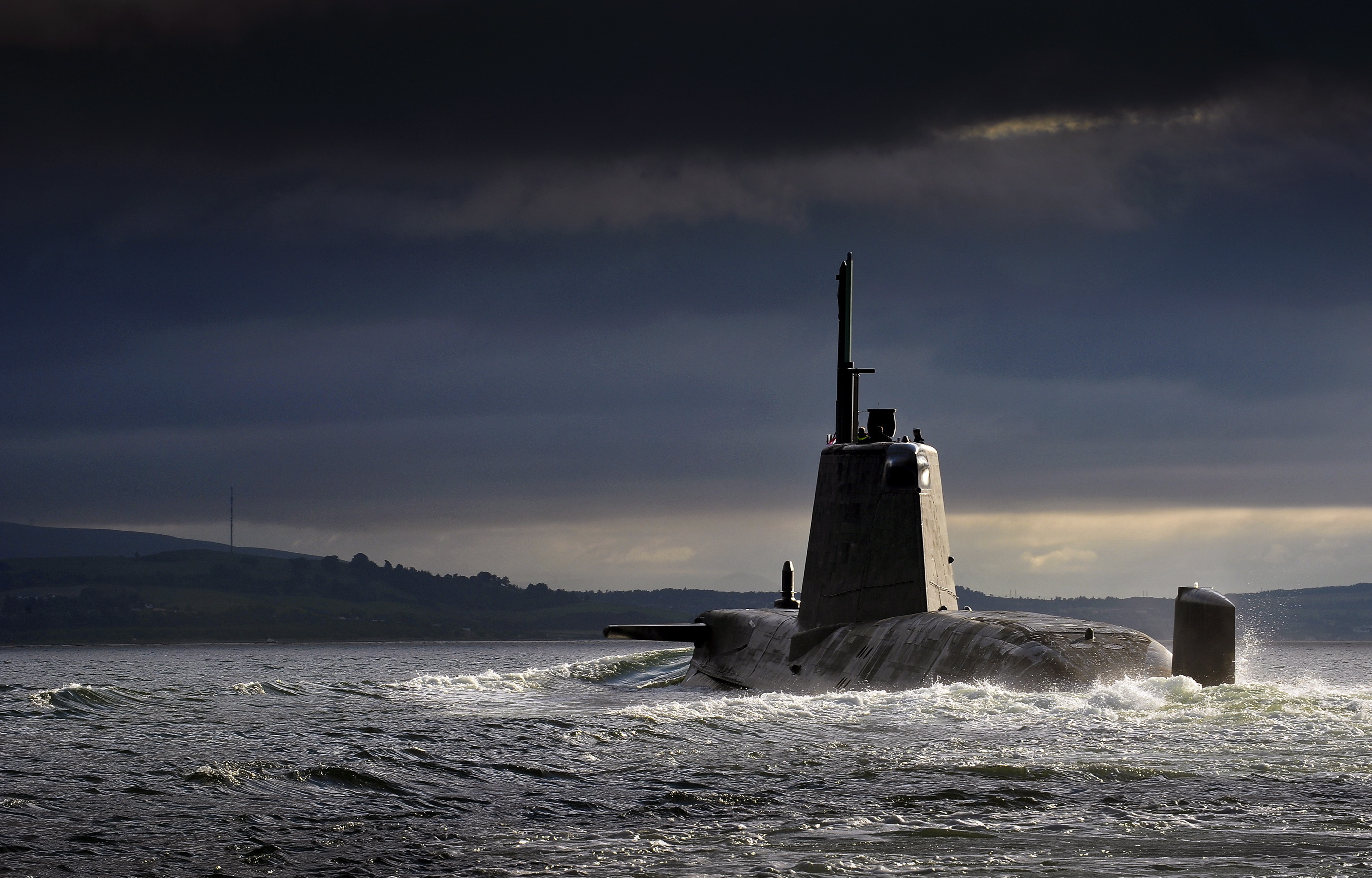 Military submarine cruising on the ocean surface under dramatic dark clouds near a distant coastline.