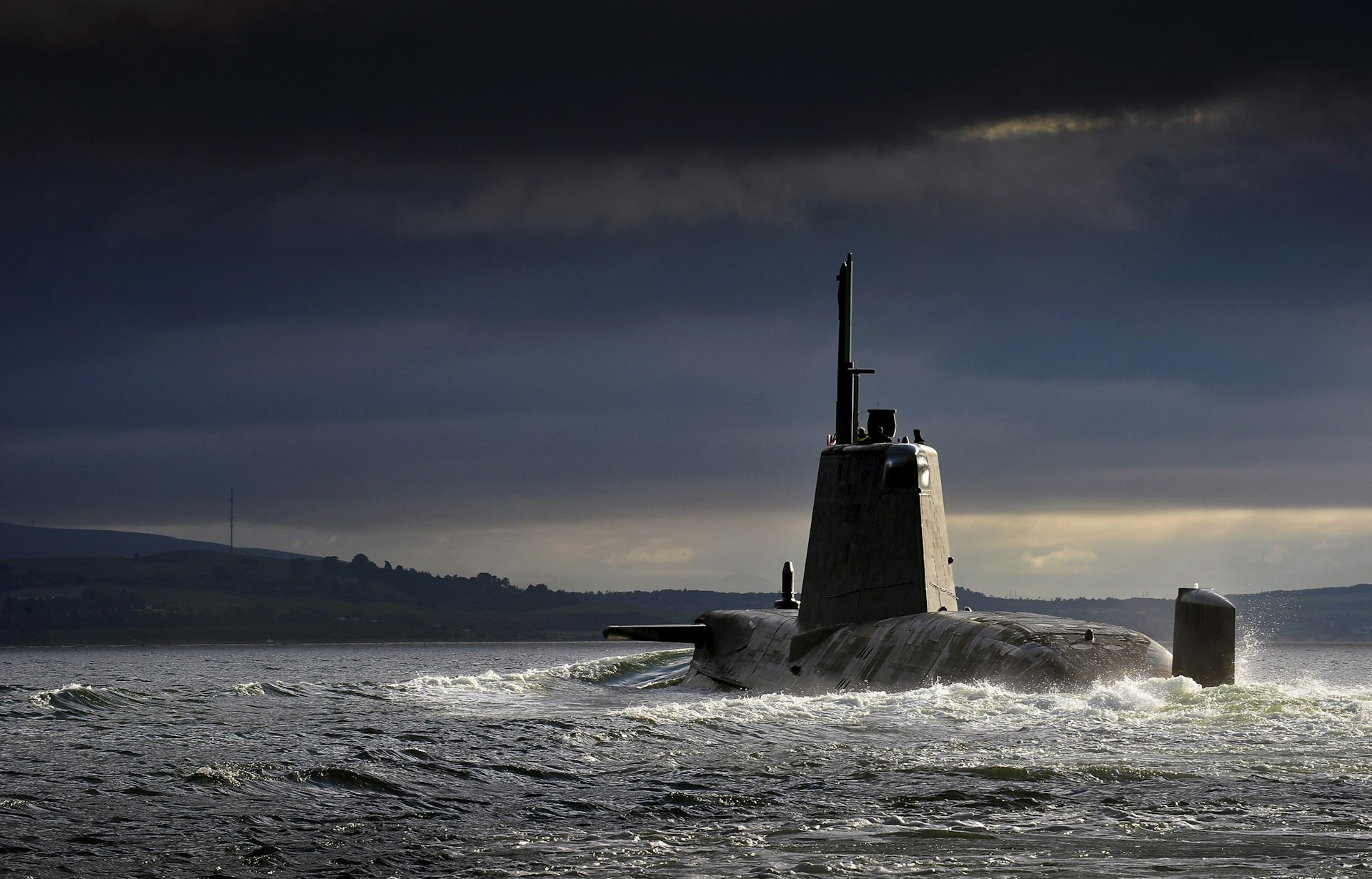Military submarine cruising on the ocean surface under dramatic dark clouds near a distant coastline.
