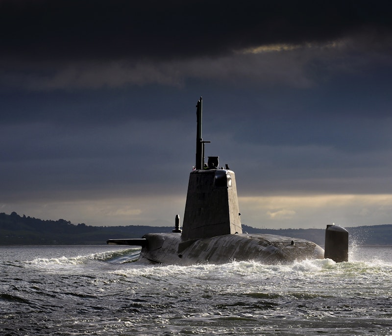 Military submarine cruising on the ocean surface under dramatic dark clouds near a distant coastline.