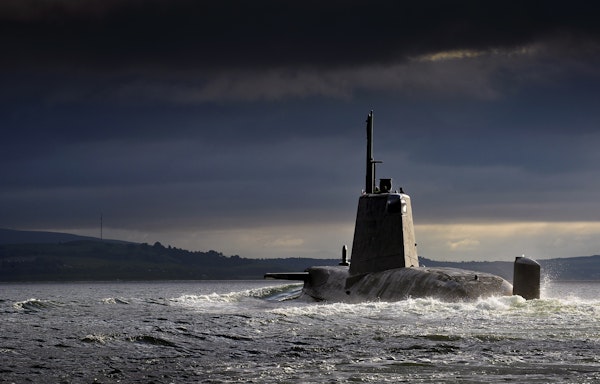Military submarine cruising on the ocean surface under dramatic dark clouds near a distant coastline.