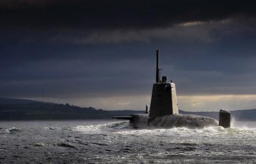 Military submarine cruising on the ocean surface under dramatic dark clouds near a distant coastline.