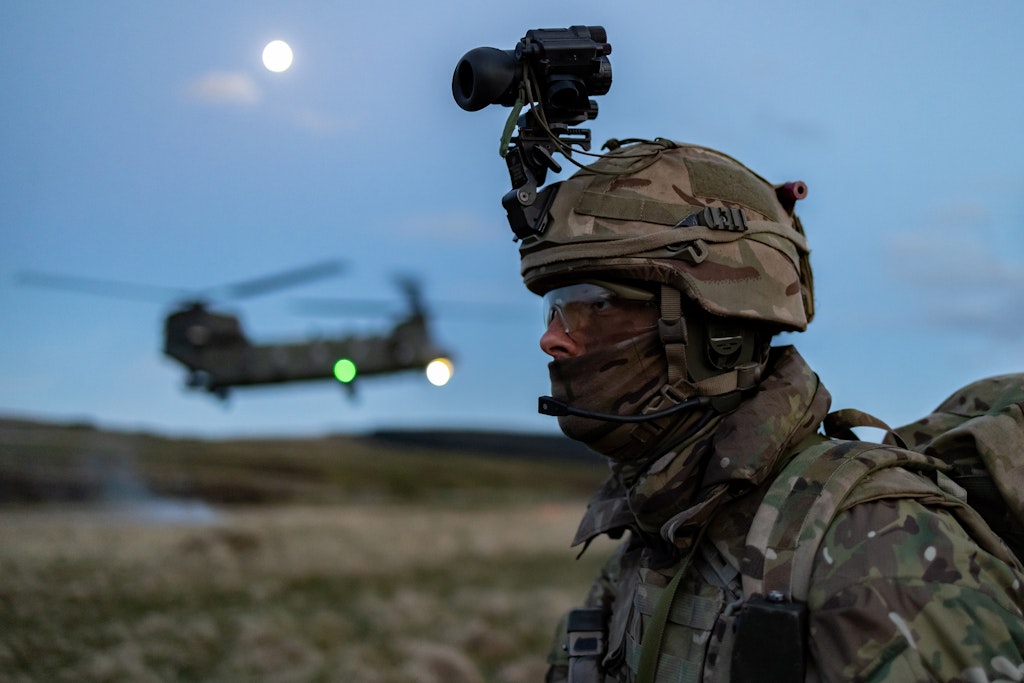 Soldier in camouflage, wearing night-vision gear, stands at dusk with a Chinook helicopter hovering in the background under a bright moon.
