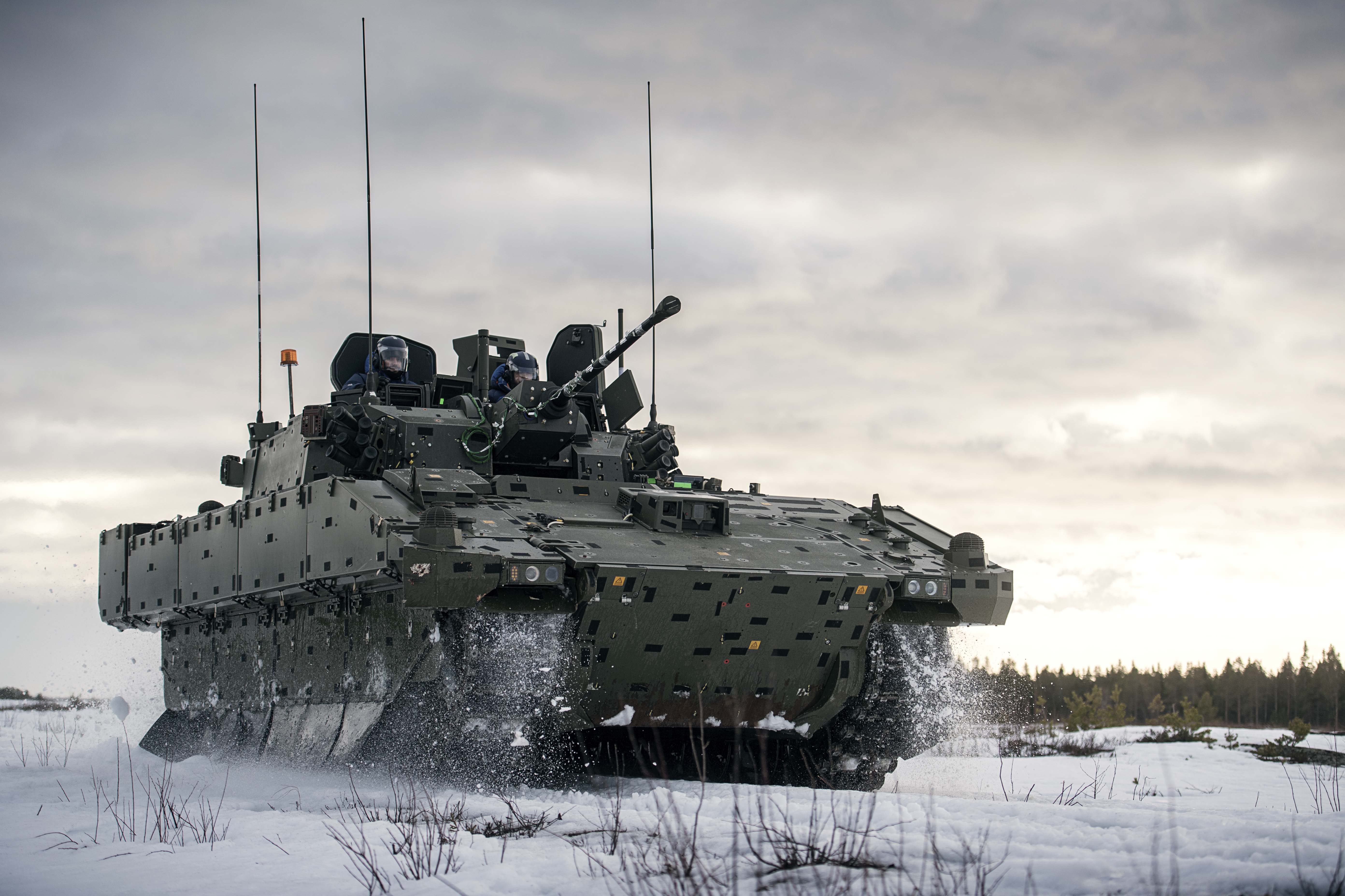 Armored military vehicle with mounted cannon driving through snow during winter military exercises.