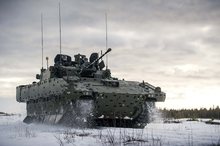 Armored military vehicle with mounted cannon driving through snow during winter military exercises.