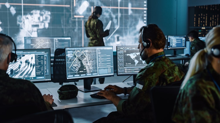Military personnel in a command center monitoring surveillance feeds, maps, and intelligence data on multiple computer screens.