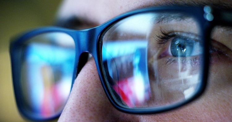 Close-up of a person’s eye with computer screen reflections visible in their glasses.