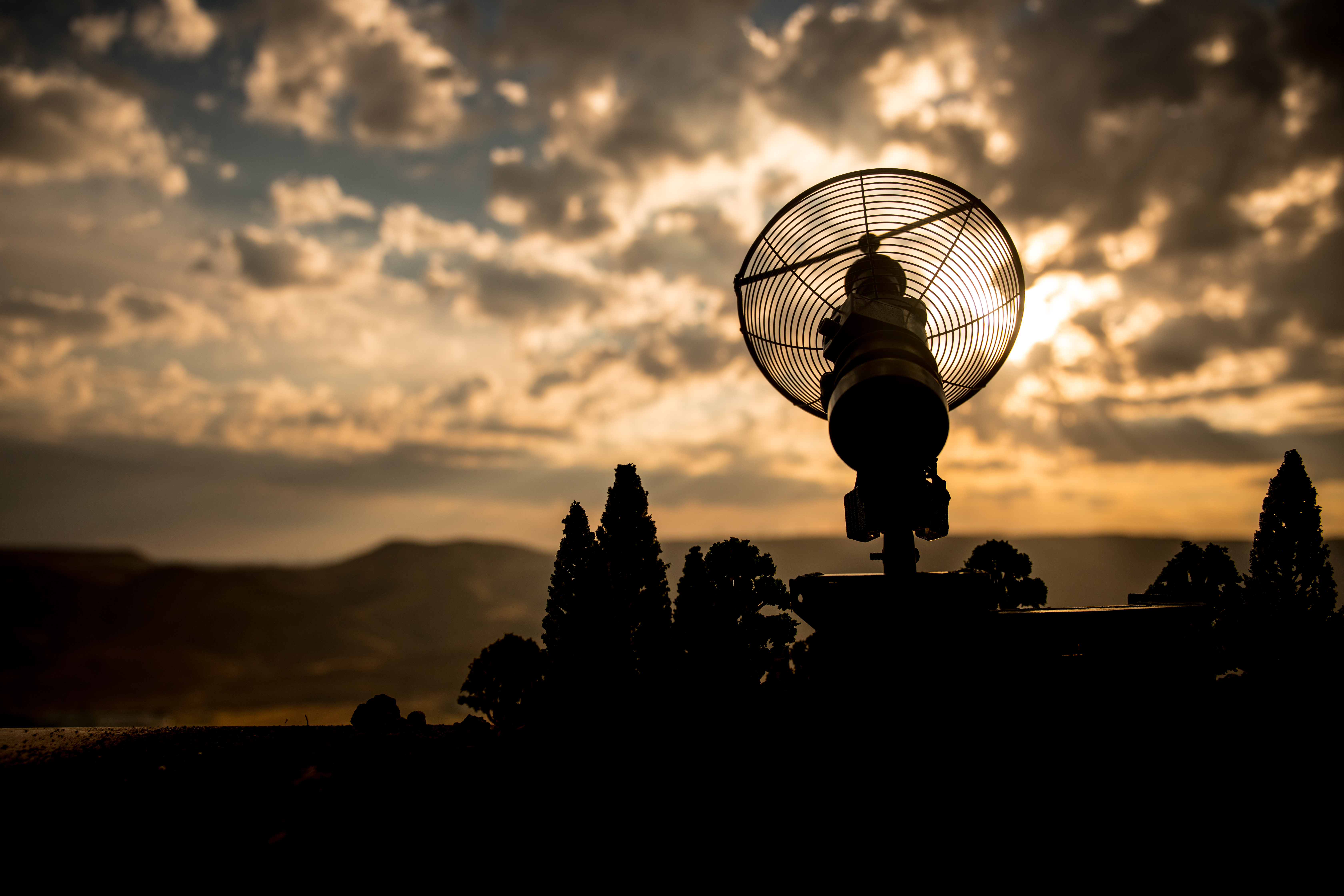 Silhouetted parabolic antenna against a glowing sunset sky with dramatic clouds and distant mountains.
