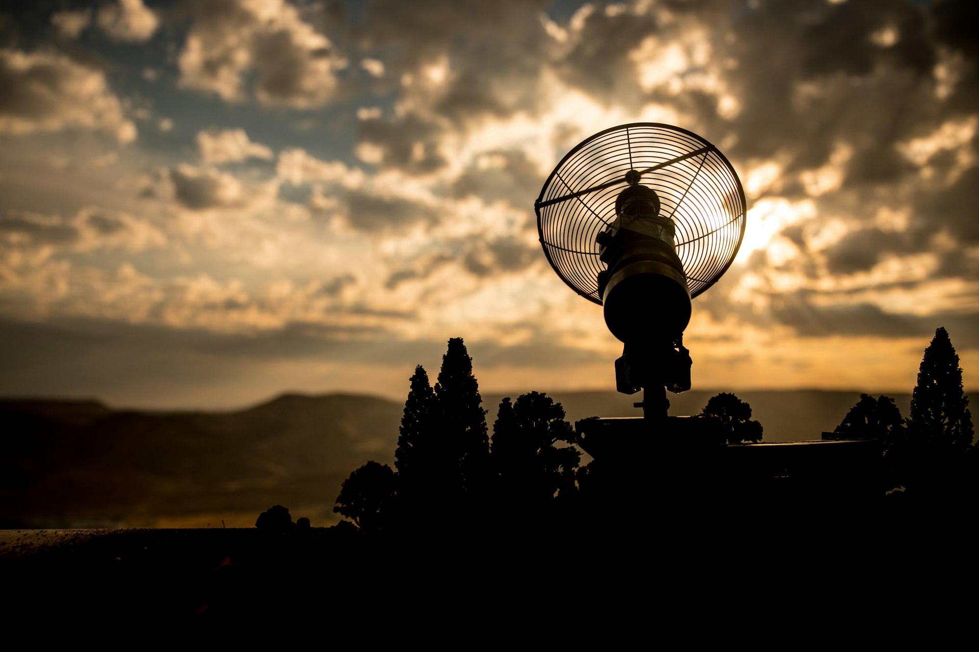 Silhouetted parabolic antenna against a glowing sunset sky with dramatic clouds and distant mountains.