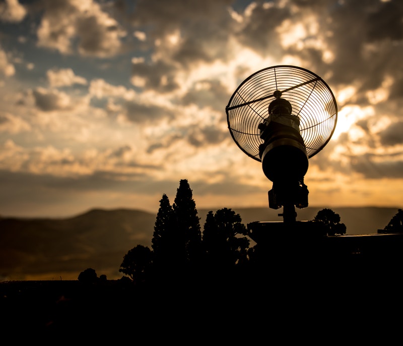 Silhouetted parabolic antenna against a glowing sunset sky with dramatic clouds and distant mountains.