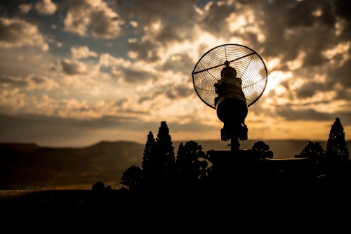Silhouetted parabolic antenna against a glowing sunset sky with dramatic clouds and distant mountains.