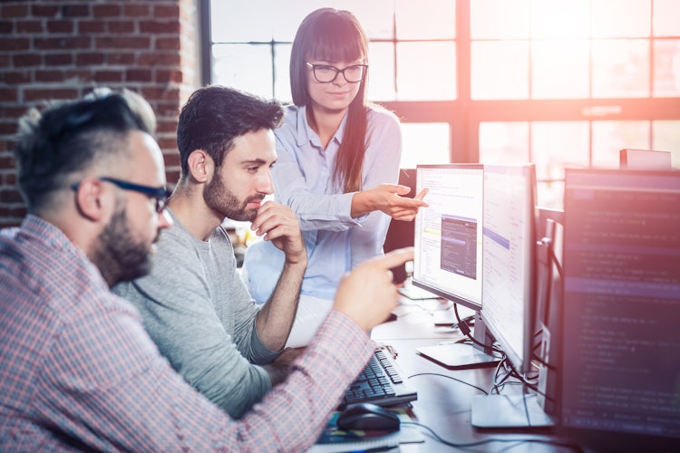 Team of software developers collaborating in a modern office, reviewing and discussing code displayed on computer monitors.